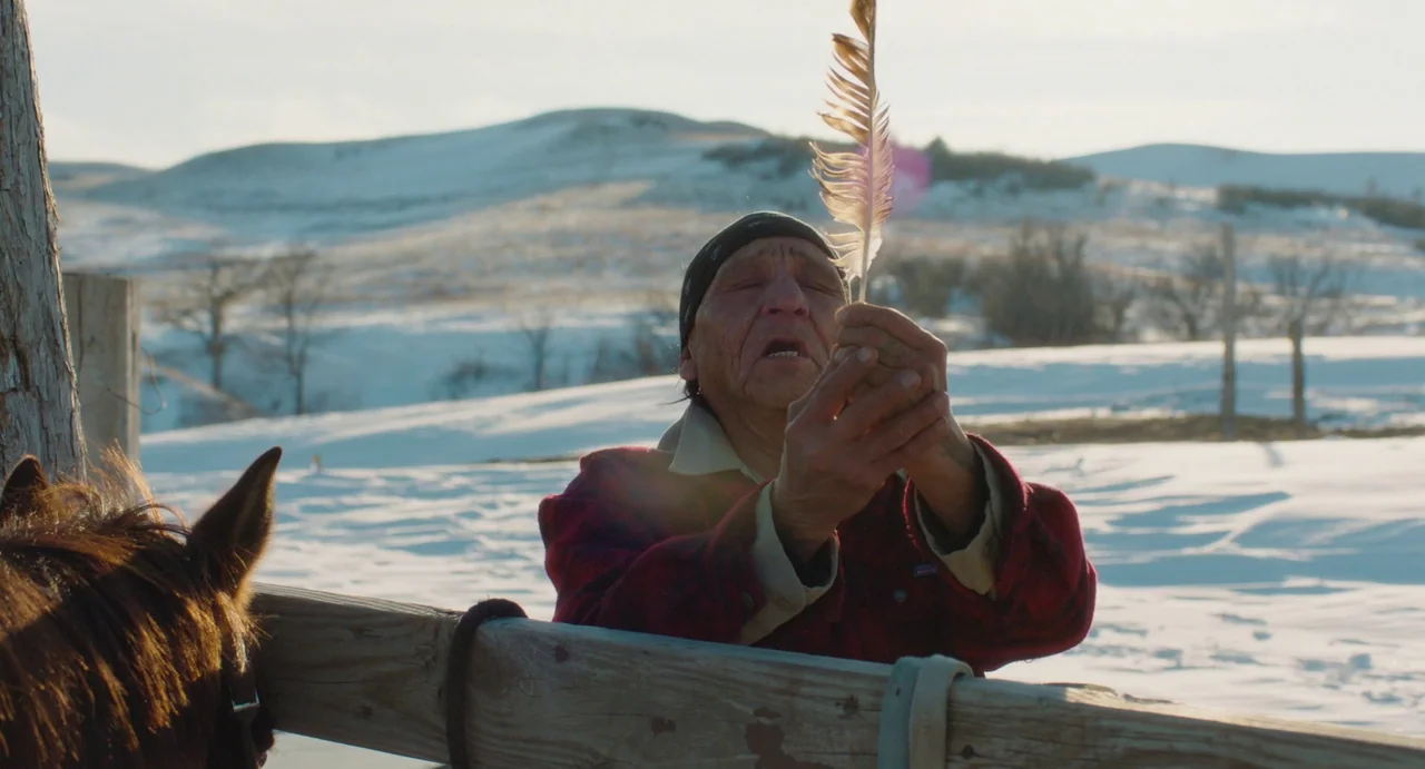 Eureka (2024) movie still: medium — A snowy field serves as the backdrop for a wooden paddock fence. An elderly man stands by the fence holding a…