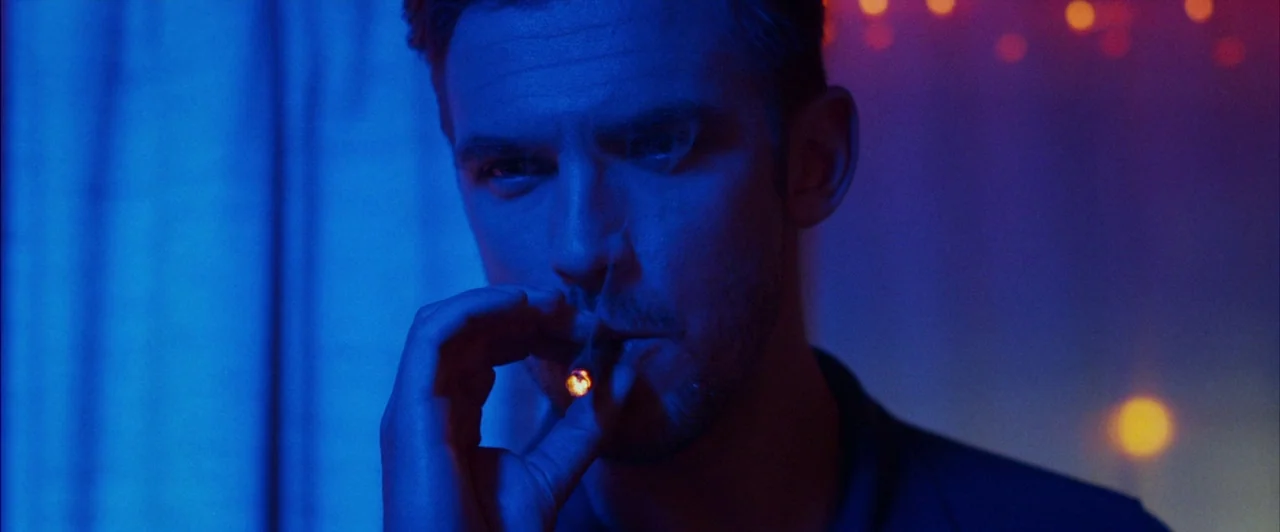 The Guest (2014) movie still: closeup — A dimly lit space features heavy blue tones and out-of-focus amber light sources behind. A man with stubble…