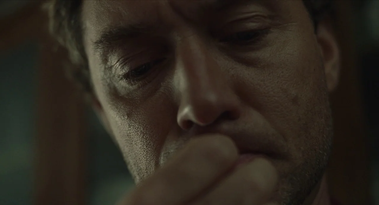 The Nest (2020) movie still: closeup — The frame shows a tight detail of a man's face in a dim space. He holds his knuckles or fingers against his…