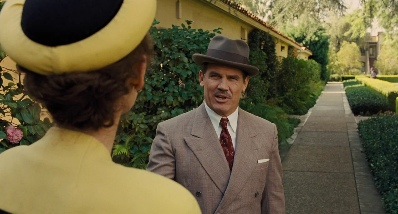 Hail, Caesar! (2016) movie still: medium — An outdoor concrete path leads past a stucco building with neatly trimmed hedges. A man in a fedora and…