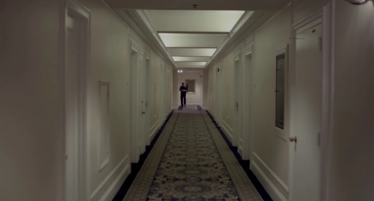 Beginners (2010) movie still: wide — A long, symmetrical hotel corridor features plain white walls with rows of closed doors. A lone man stands in…