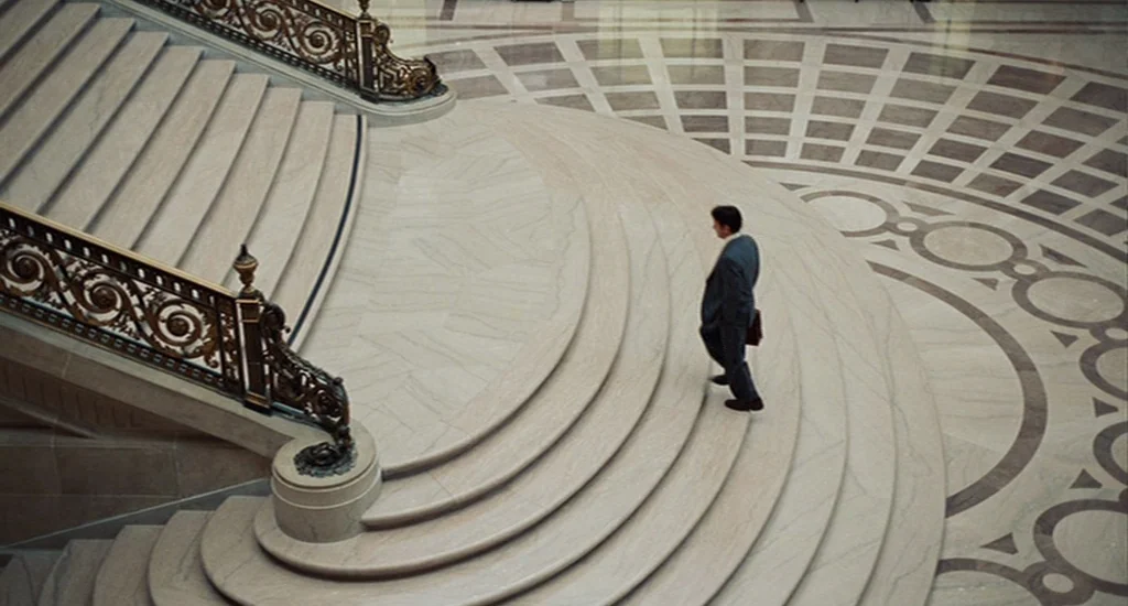 Milk (2008) movie still: wide — A spacious marble lobby features a grand curved staircase with ornate dark metal railings. A solitary businessman in…