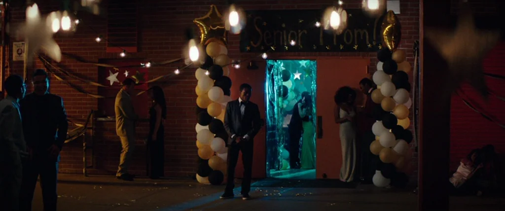 Dope (2015) movie still: wide — An outdoor brick wall area is decorated for a high school senior prom event. Teenagers in formal attire gather near…