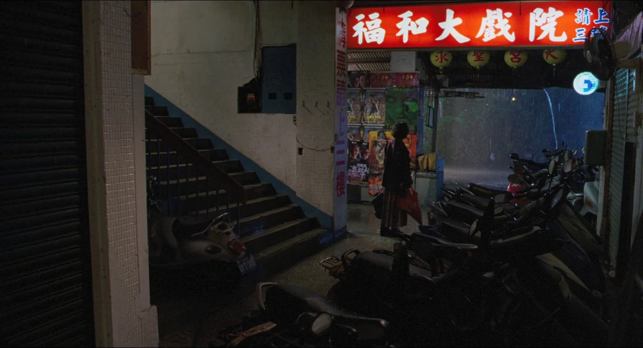 Goodbye, Dragon Inn (2003) movie still: wide — A dimly lit covered walkway outside a theater features a vibrant red neon sign. A woman stands alone…