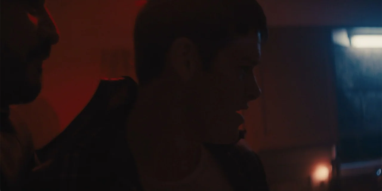 All Roads to Pearla (2019) movie still: medium — A dimly lit interior space features high-contrast red and blue light sources. Two men are…