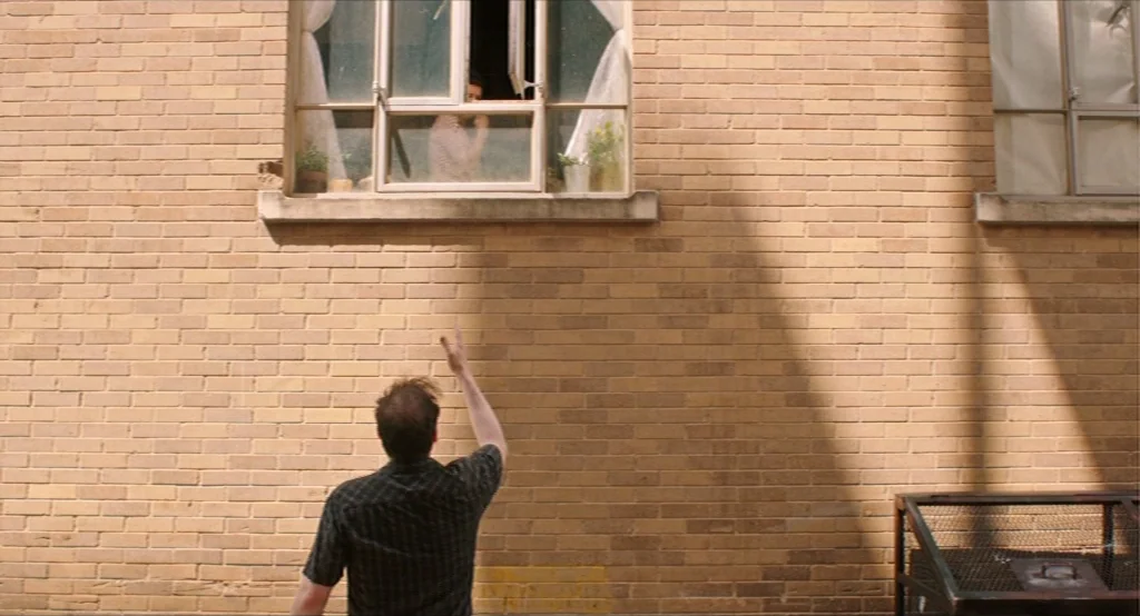 Results (2015) movie still: medium — An exterior brick building wall features two rectangular windows with thin white curtains. A man stands in…
