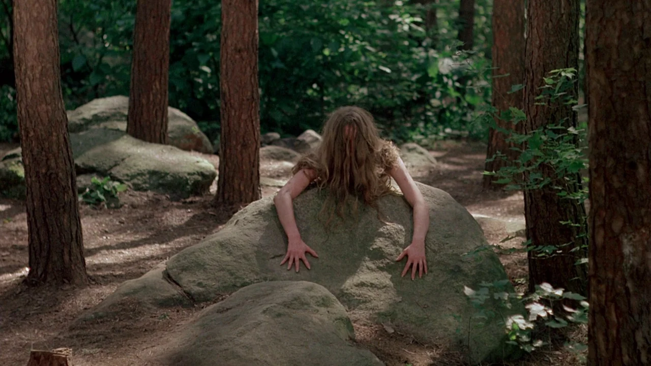 I Spit On Your Grave (1978) movie still: medium — A dense forest floor features tall pine trees and moss-covered granite boulders. A woman lies flat…