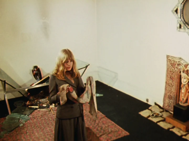 Lucifer Rising (1972) movie still: medium — An interior space with white walls and a dark carpet features scattered broken glass. A woman in a grey…