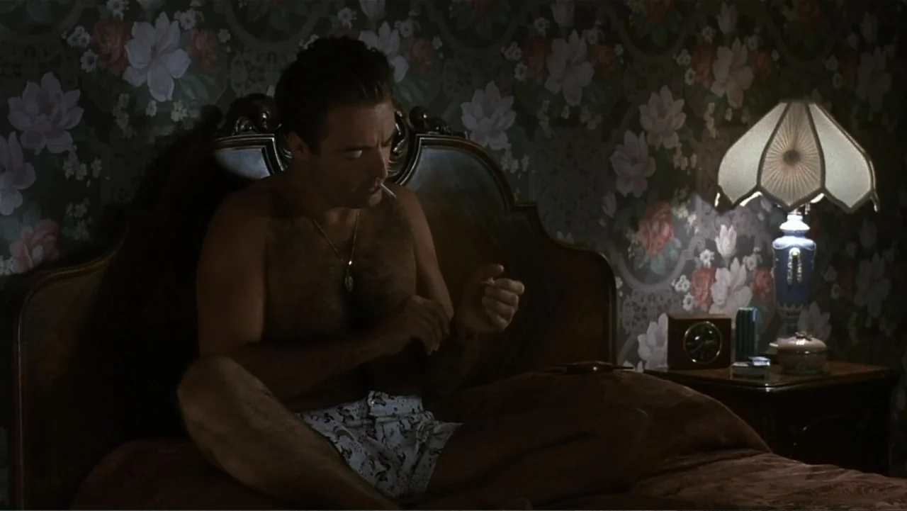 The Mambo Kings (1992) movie still: medium — A bedroom features floral wallpaper and a dark wood headboard illuminated by a lamp. A man sits…