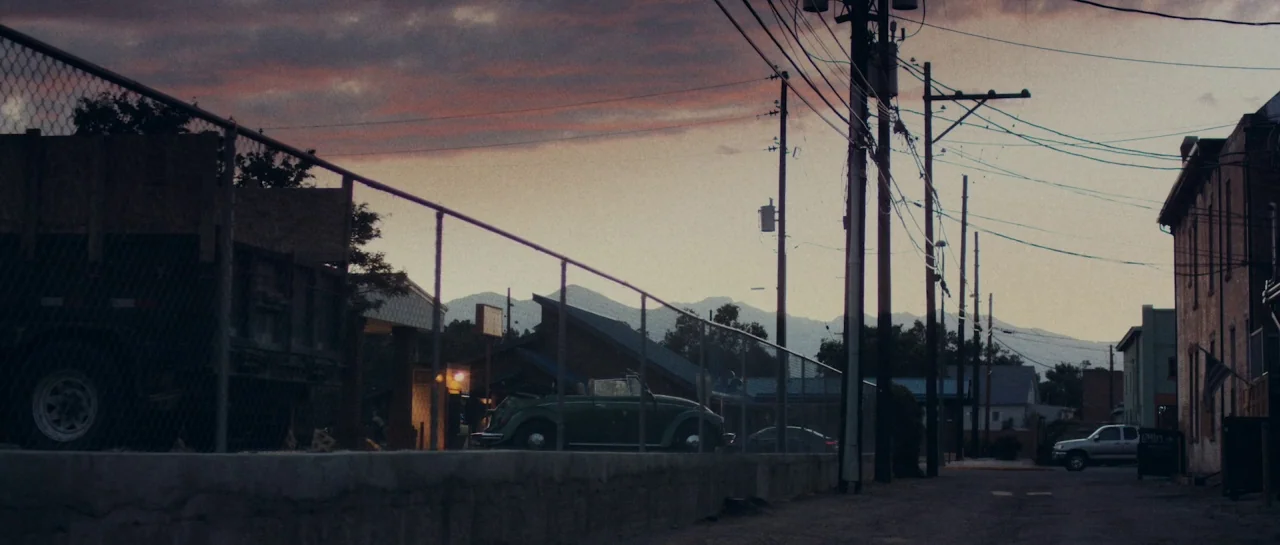Starfish (2018) movie still: establishing — A quiet, paved street runs past a chain-link fence and a small green car. Distant mountains frame the…