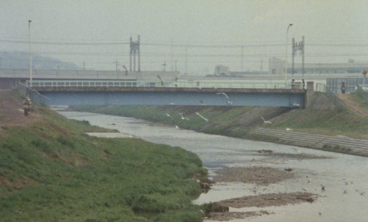 Evil Dead Trap (1988) movie still: establishing — A wide, shallow river flows under a simple blue steel bridge in a rural landscape. Grassy banks…