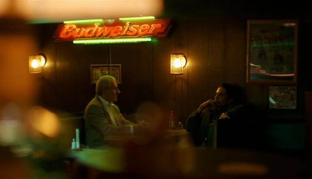 The Limey (1999) movie still: wide — A dark, wood-paneled corner of a restaurant features a backlit Budweiser neon sign. Two men sit in a vinyl…
