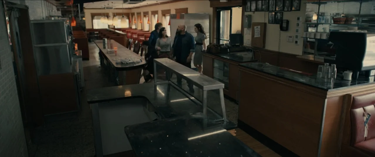 Guns Up (2025) movie still: wide — A quiet diner interior with red vinyl booths and wood-paneled walls. Four adults stand in the walkway between the…