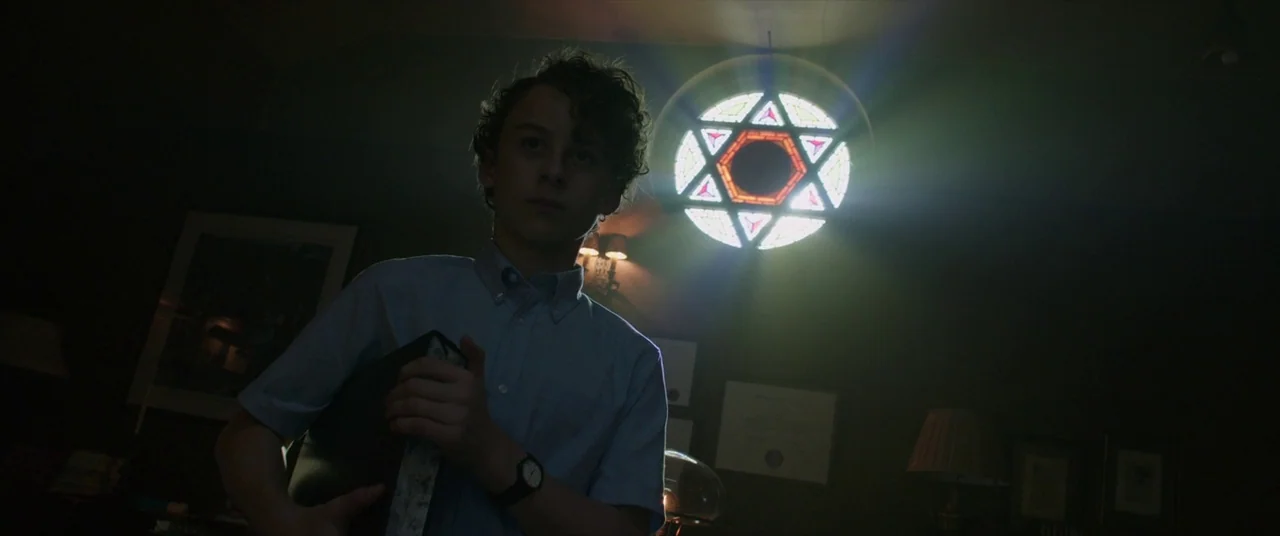 It (2017) movie still: medium — A dimly lit room features a prominent circular stained glass window depicting a hexagram. A teenage boy holds a…