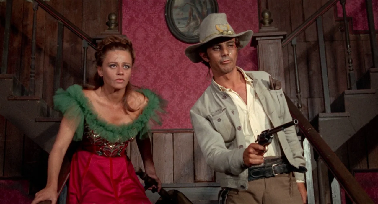 The Hellbenders (1967) movie still: medium — A woman and a man in Western-style clothing stand on a wooden staircase with red wallpaper. The man…