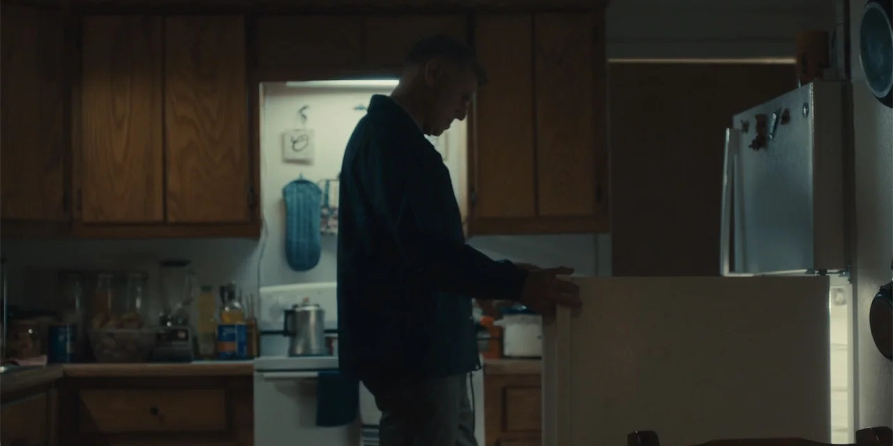 All Roads to Pearla (2019) movie still: medium — A dimly lit room features wood cabinetry and a humming, open refrigerator at night. A middle-aged…