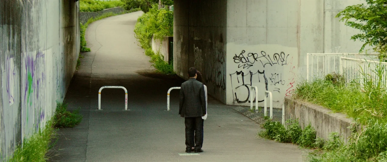 Creepy (2016) movie still: wide — A paved path runs beneath a concrete bridge structure covered in faded graffiti tags. Two people stand in the…