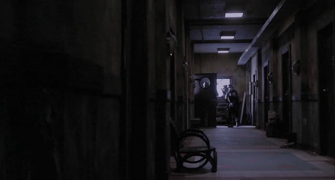 The Raid (2012) movie still: wide — A dimly lit narrow hallway with darkened doors and stained, worn tile flooring. Two figures in tactical gear…