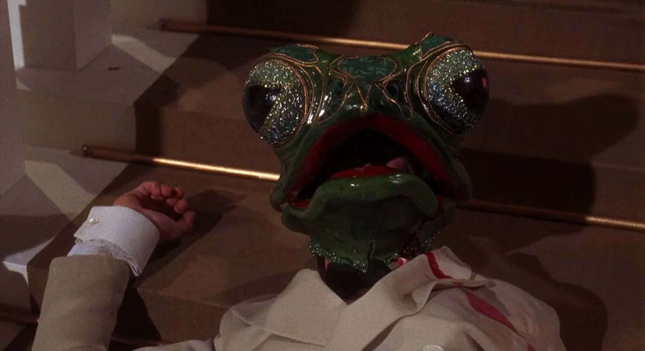 The Abominible Dr. Phibes (1972) movie still: medium — A person in a formal suit lies across wooden stairs wearing a large mask. The frog-shaped…
