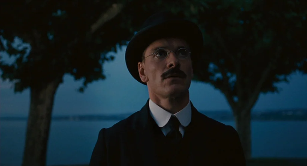 A Dangerous Method movie still: medium — A man stands outdoors near a large body of water during twilight. Trees are visible in the blurry…