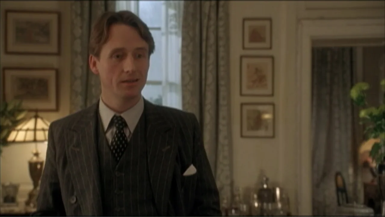 The Gathering Storm (2002) movie still: medium — A traditional room features framed pictures on the wall and curtains by the window. A man in a…