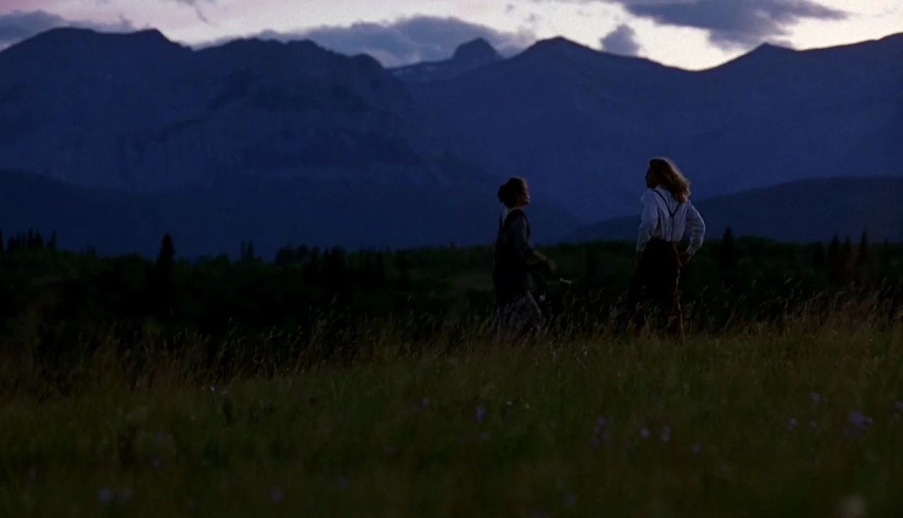 Legends of the Fall (1994) movie still: wide — A high mountain range stands behind a vast, dark field under a twilight sky. Two people stand facing…