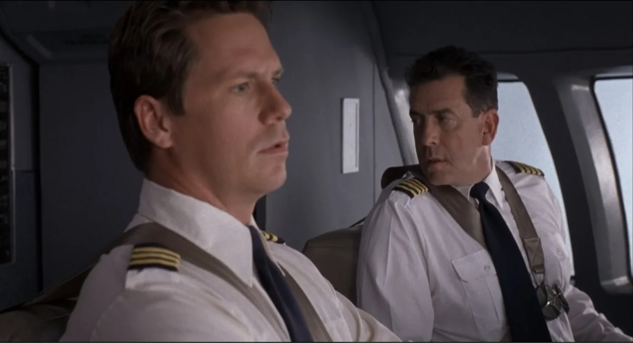 Air Rage (2001) movie still: medium — A cramped airplane cockpit features dark gray walls and large windows looking outward. Two male pilots wearing…