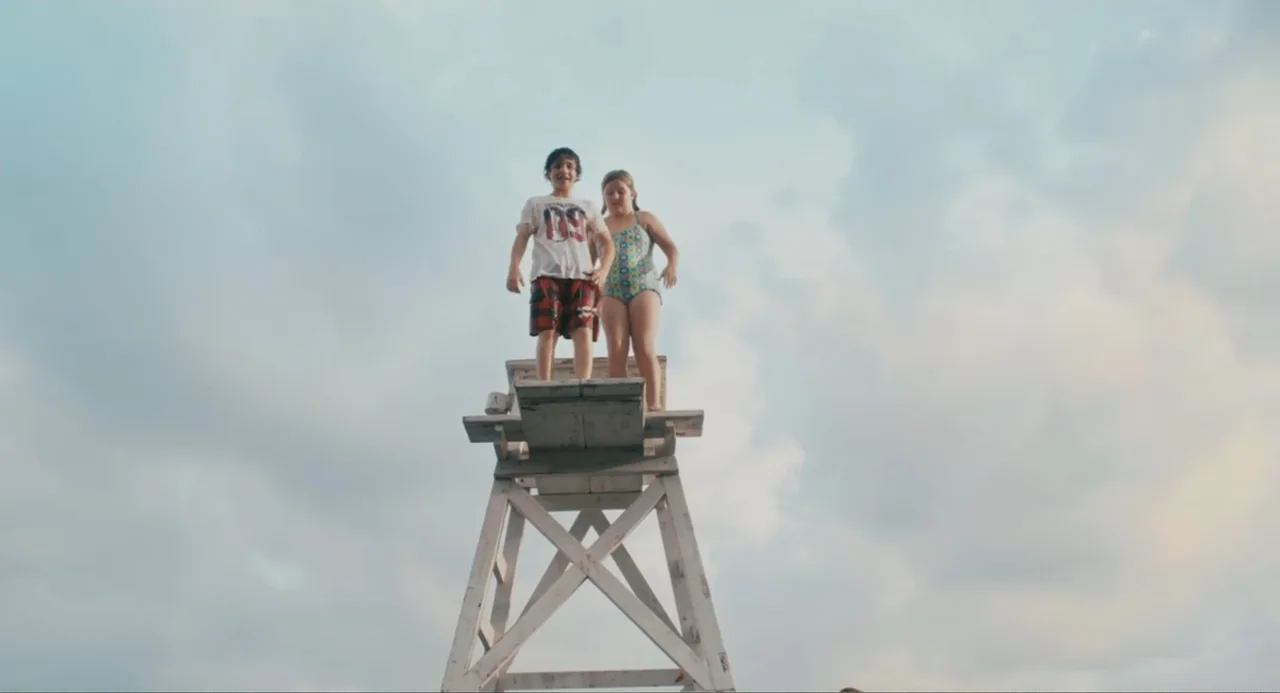 Grown Ups (2010) movie still: fullbody — A tall white wooden lifeguard chair sits against a bright overcast sky. A young boy and girl stand together…