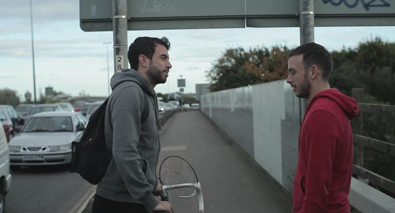 Weekend (2011) movie still: medium — A concrete pedestrian overpass overlooks a multi-lane roadway with moving cars. Two men converse near a metal…