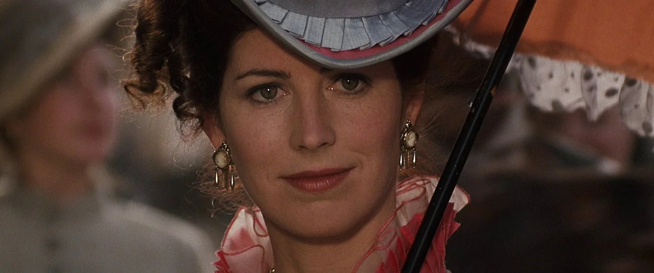 Tombstone (1993) movie still: closeup — An outdoor setting with blurred figures suggests a public event or park gathering. A woman in period…