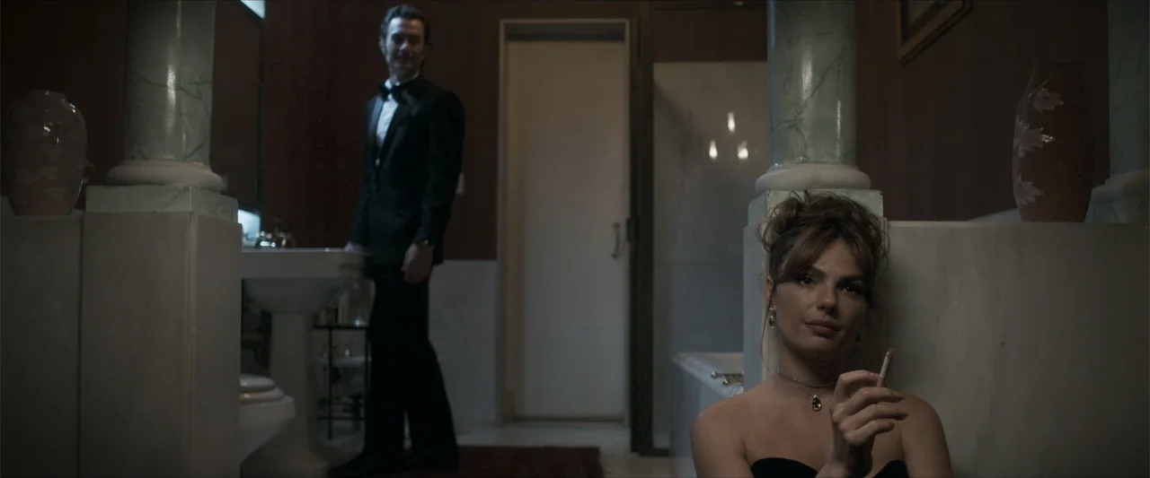 Angela (2023) movie still: medium — A dark, elegant bathroom features marble-like columns and a door in the center background. A woman in a dress…
