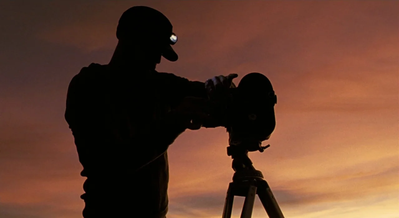 Patagonia (2010) movie still: medium — A person stands outdoors against a sunset sky with clouds. They adjust a professional camera mounted on a…