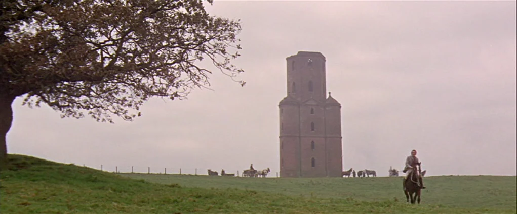 Far from the Madding Crowd (1967) movie still: wide — An open green pasture under an overcast sky features a tall brick tower. A man rides a horse…