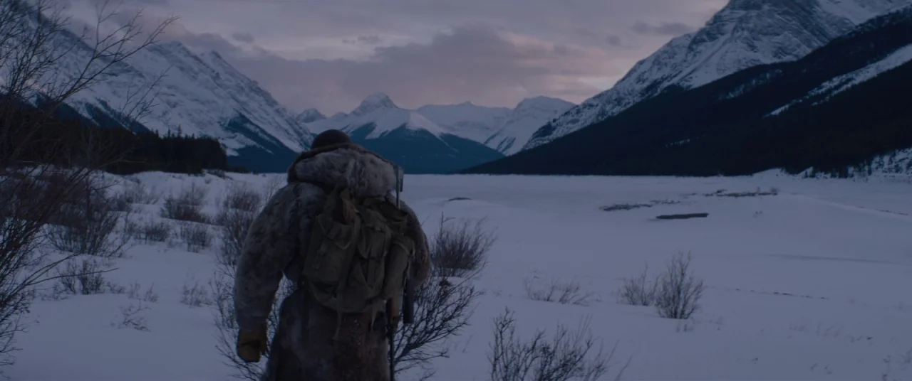 Hold The Dark (2018) movie still: fullbody — A snow-covered mountain valley stretches toward a massive frozen lake at twilight. A solitary man in a…