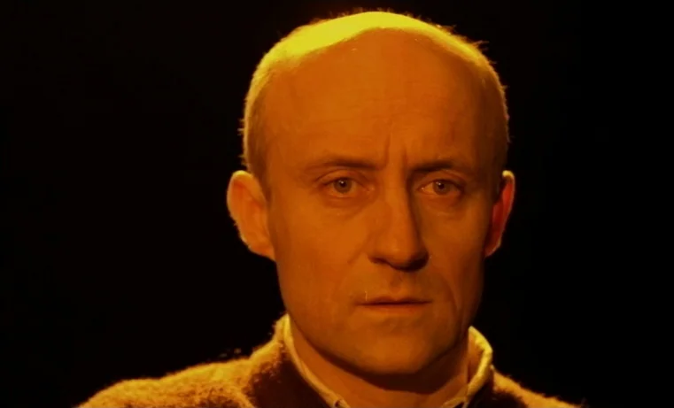 Golem (1980) movie still: closeup — A man stands in a dark environment with a plain black background. He looks ahead with a steady, fixed gaze.