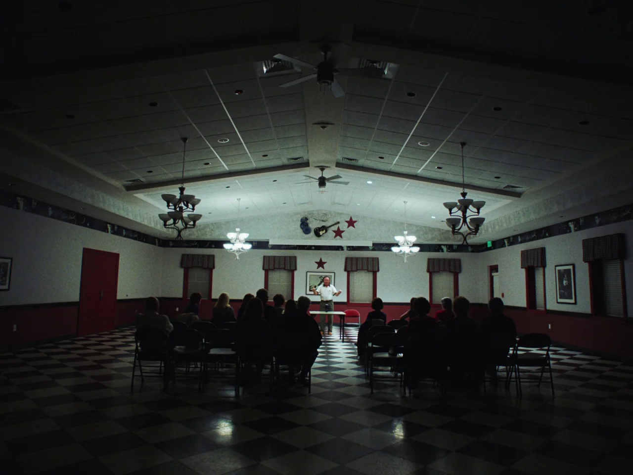 Some Kind Of Heaven (2020) movie still: wide — A wide community room features a black and white checkered tile floor and white walls. A small group…