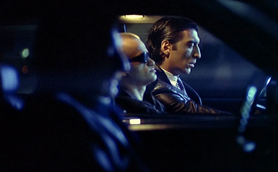 Diva (1981) movie still: medium — The interior of a moving car at night is dimly lit by passing streetlights. Two men sit in the front seats looking…