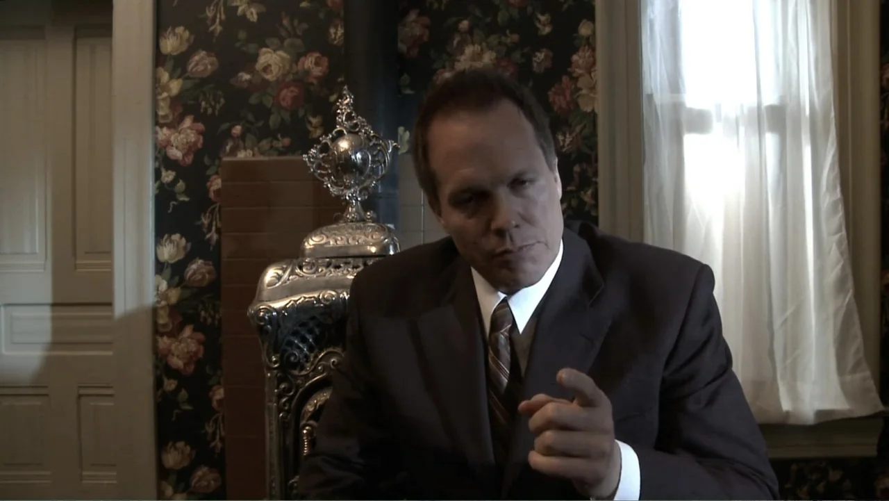 Boathouse Detectives (2010) movie still: medium — A room with vintage floral wallpaper features a detailed silver wood-burning stove behind a man.…