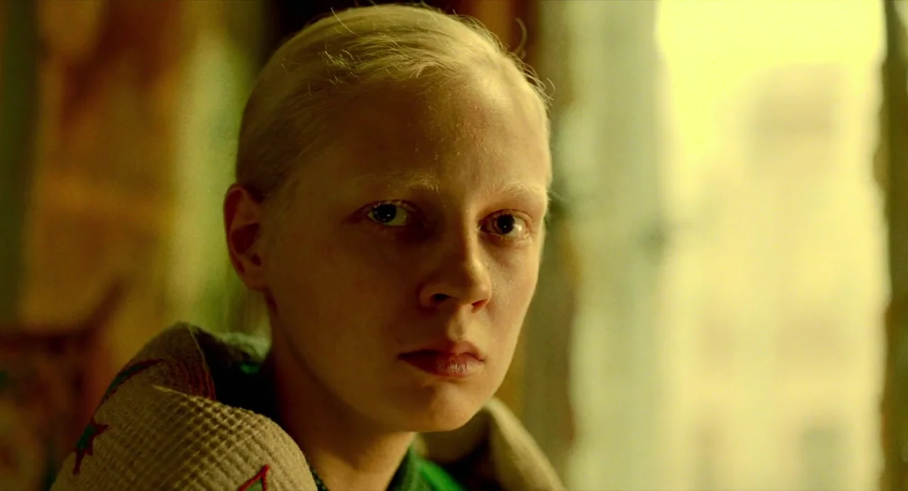 Beanpole (2019) movie still: closeup — A person with pale features is indoors next to a bright window source. They gaze directly into the lens while…