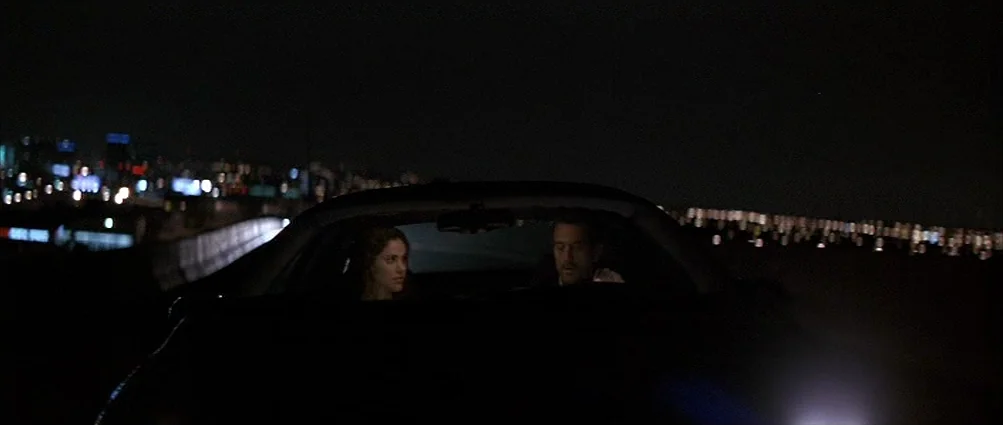 Heat (1995) movie still: wide — A man and woman sit inside a dark car driving at night on a highway. The glowing city skyline is visible in the…