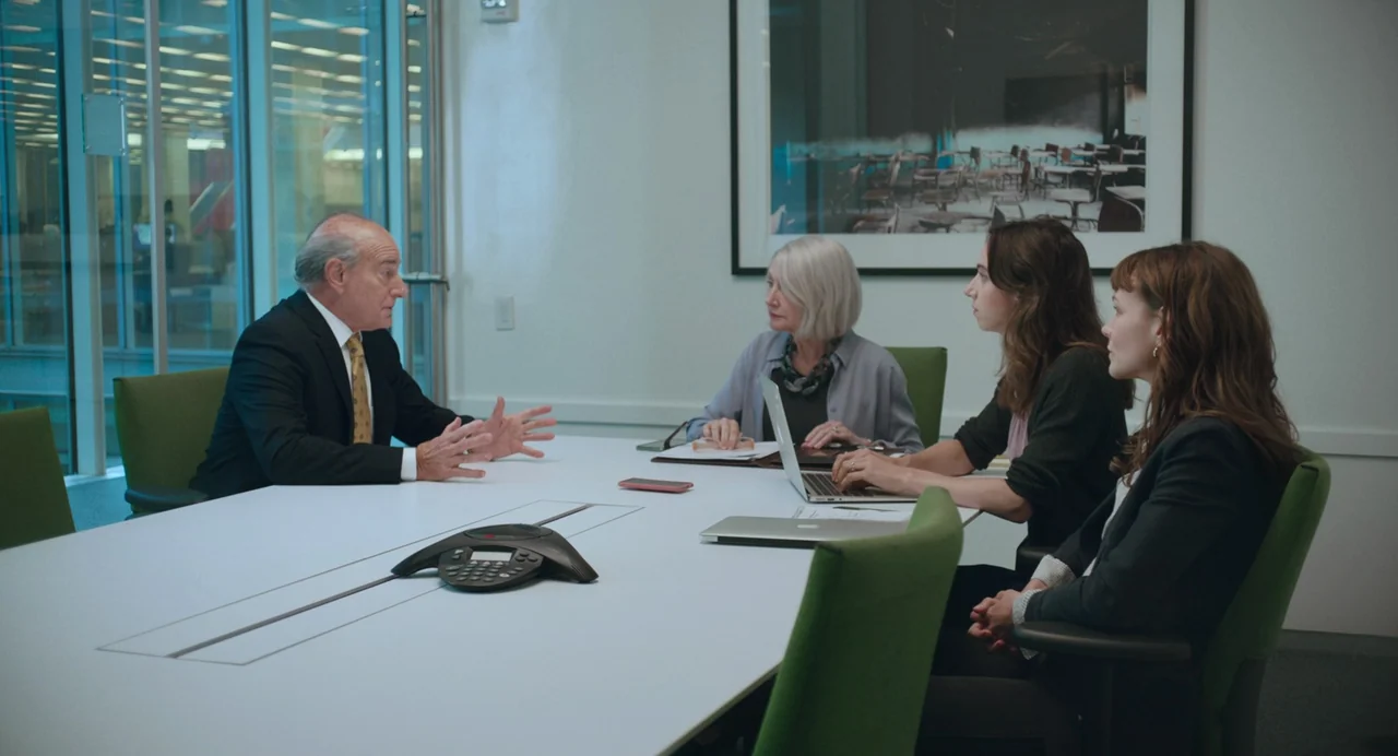 She Said (2022) movie still: wide — A modern, bright conference room features a long white table and green chairs. Four professionals sit together…