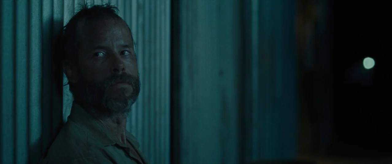 The Rover (2014) movie still: medium — A dark, narrow outdoor corridor features a vertical metal corrugated wall surface. A bearded man leans…