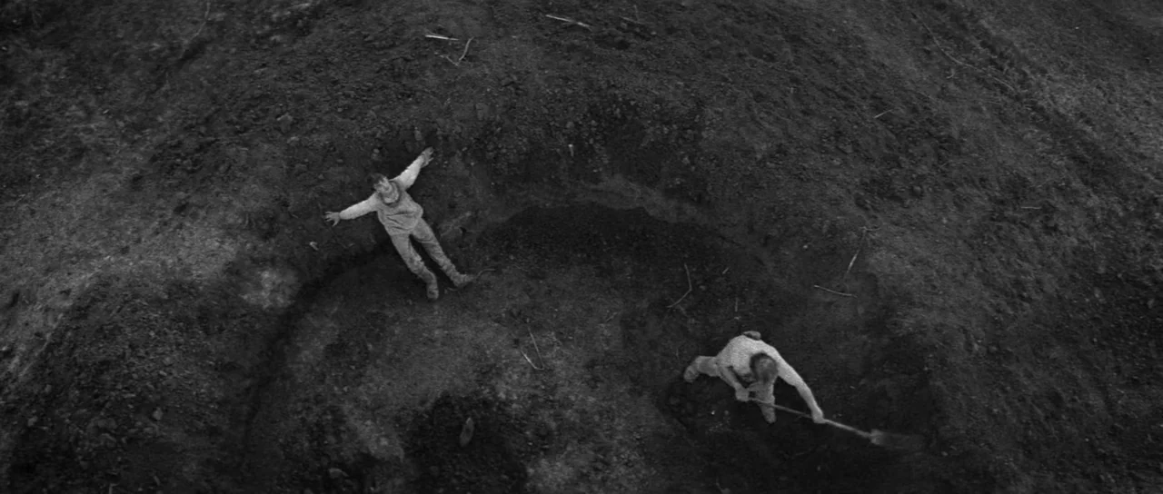 Andrei Rublev (1966) movie still: wide — A deep circular pit carved into raw dark earth under an overcast sky. One man lies on his back with arms…