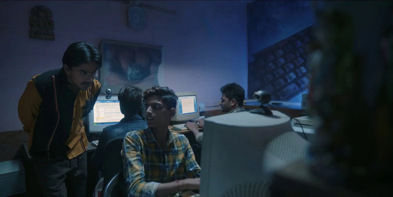 The White Tiger (2021) movie still: medium — A dimly lit room contains multiple CRT computer setups arranged on tables. A man stands behind another…