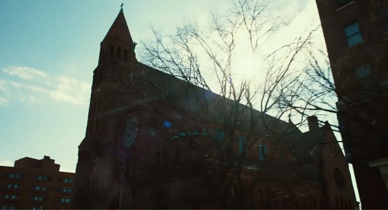 An Imperfect Murder (2017) movie still: establishing — A large brick church building stands under a bright clear sky during the day. Bare branches…
