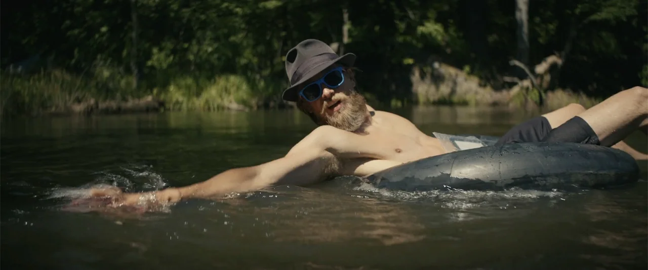 Last Looks (2022) movie still: medium — A man floats in a calm river surrounded by dense green forest trees. He wears a hat and sunglasses while…