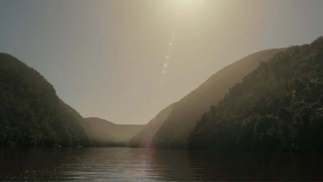 The Dinosaur Project (2012) movie still: establishing — A wide river winds between two large hills covered in dense green vegetation. Sunlight glows…