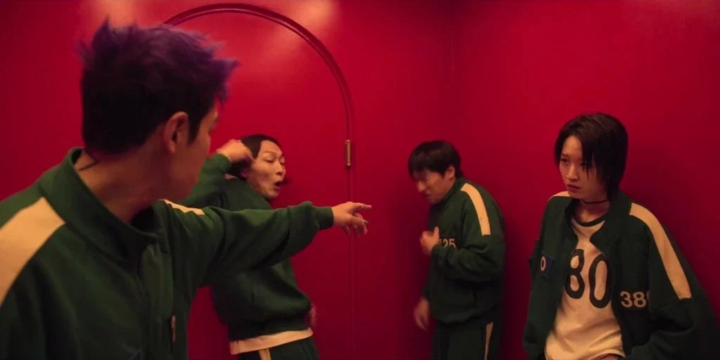 Squid Game (2024) movie still: medium — A windowless room features bright red walls and an arched doorway outline. Four individuals wearing matching…