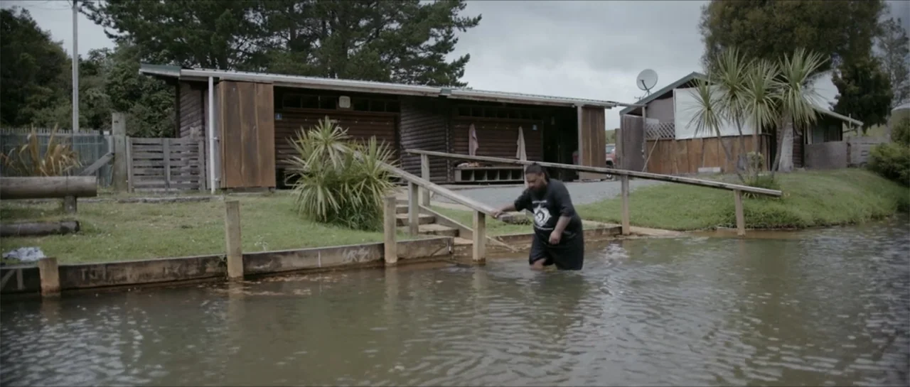 Bludgeon (2018) movie still: wide — A man in black clothing wades through murky water toward a wooden boat ramp. A simple wooden cabin with a…