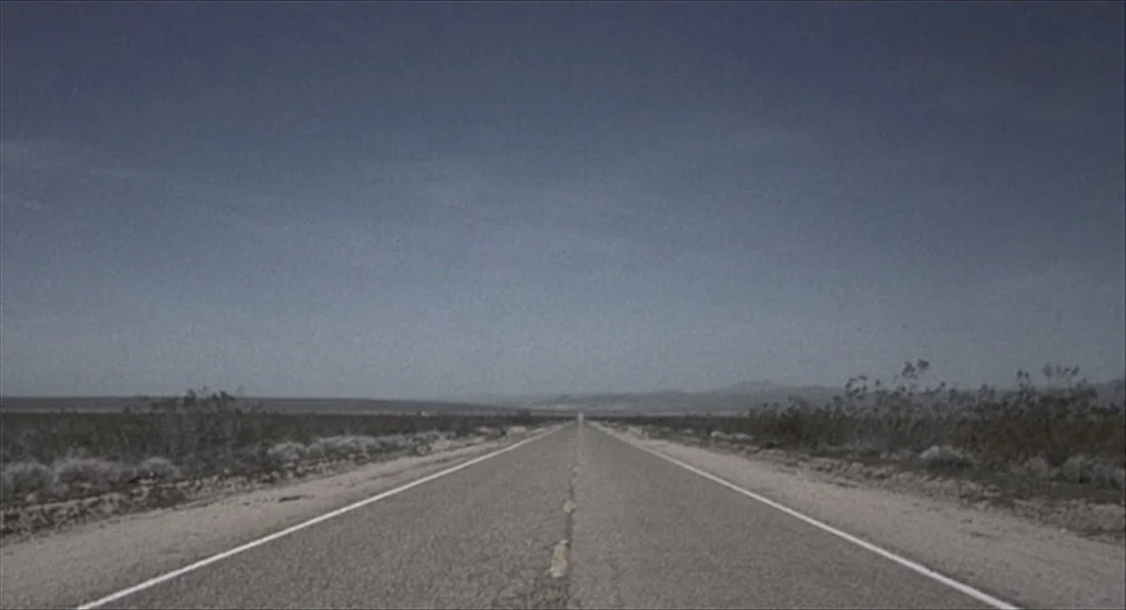 The Good Girl (2002) movie still: establishing — A long, straight asphalt road bisects a vast, flat desert landscape under clear skies. Sparse dry…